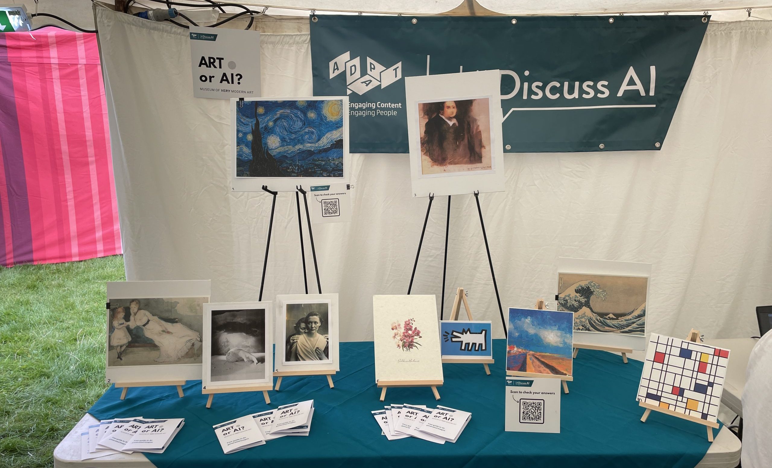 A table with a teal green tablecloth. On the table there are a number of artworks displlayed on easels. Behind the table, there is a banner with the ADAPT logo and the #DiscussAI logo.