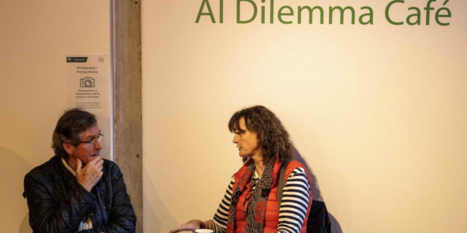 Two people are sitting and having a discussion at the "AI Dilemma Café." at European Researchers' Night 2023 at the Douglas Hyde Gallery, Trinity College Dublin. The person on the left is an older man with glasses, wearing a dark jacket and a thoughtful expression, with his hand near his chin. The person on the right is a woman with curly hair, dressed in a red vest over a striped shirt, gesturing with her hands as she speaks. There is a sign on the wall that mentions photography and videography at the event.