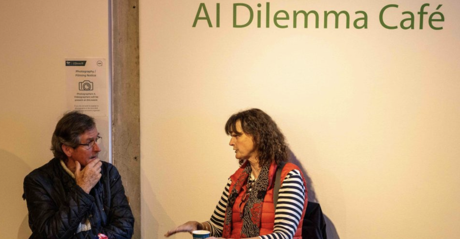 Two people are sitting and having a discussion at the "AI Dilemma Café." at European Researchers' Night 2023 at the Douglas Hyde Gallery, Trinity College Dublin. The person on the left is an older man with glasses, wearing a dark jacket and a thoughtful expression, with his hand near his chin. The person on the right is a woman with curly hair, dressed in a red vest over a striped shirt, gesturing with her hands as she speaks. There is a sign on the wall that mentions photography and videography at the event.