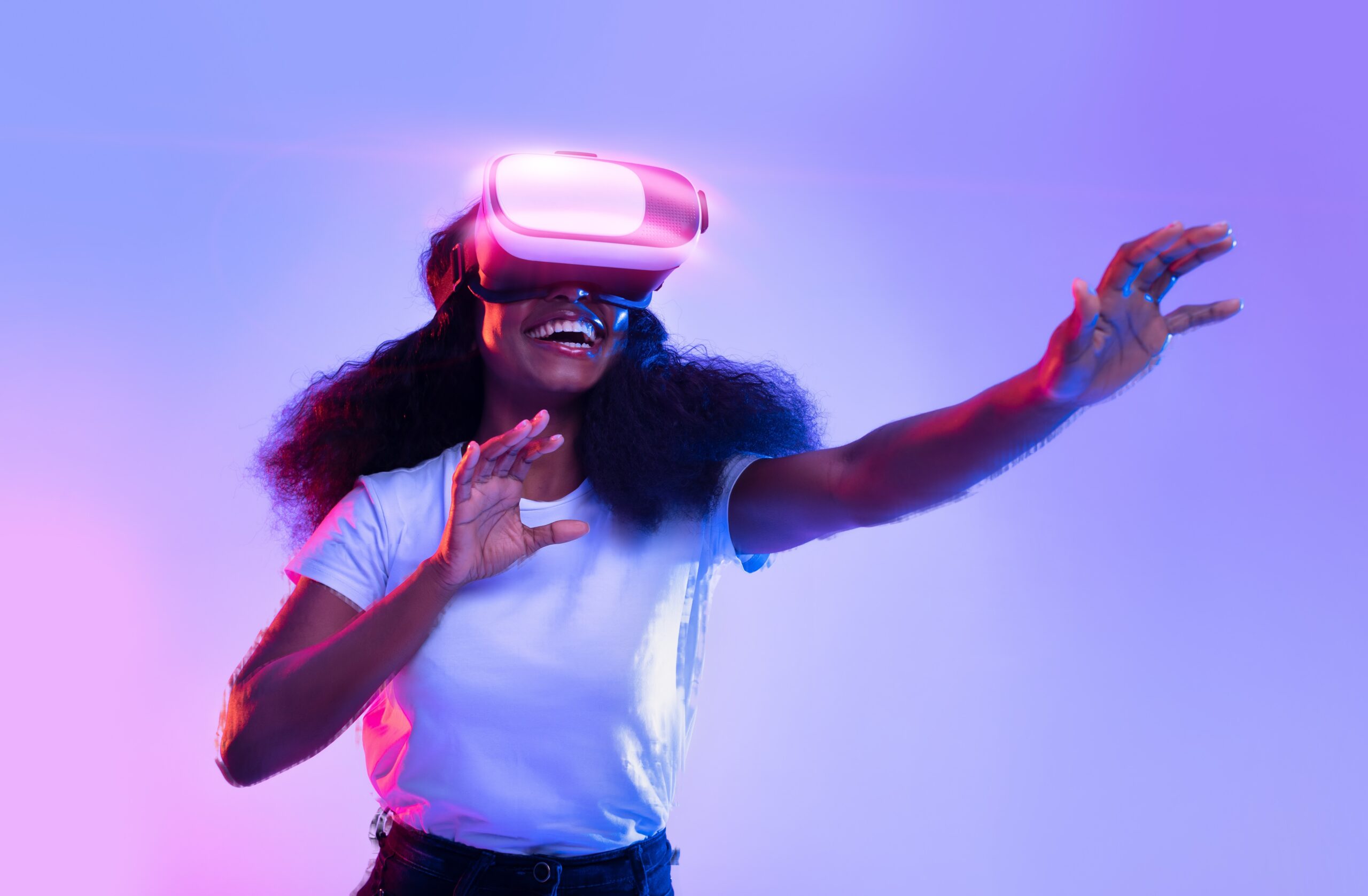 A young woman wearing a white t-shirt is experiencing virtual reality with a VR headset. She is smiling broadly, with one hand raised as if interacting with a virtual environment. The background is illuminated with a gradient of blue and purple light, giving the scene a futuristic and vibrant feel.