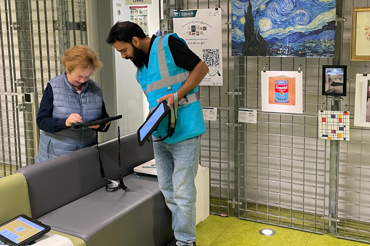 two individuals interacting with tablet devices in front of an art exhibit. The person on the left, an older woman, is focused on her tablet, while the person on the right, wearing a bright blue reflective vest, is assisting her with another tablet. The background features a display of famous artworks, including Van Gogh’s Starry Night, Andy Warhol’s Campbell’s Soup Cans, and a Mondrian-inspired piece, mounted on a metal grid. A sign on the wall references "Is it Art or AI?" and includes a QR code for further interaction, indicating that the exhibit explores AI-generated art versus traditional art.