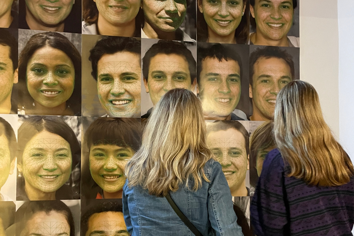 two women standing in front of a large wall display featuring multiple portrait photographs. Each portrait depicts a person's face with green geometric facial recognition mapping overlaid on the image, indicating the use of technology, likely AI, to analyze or track facial features. The women are observing the images closely, with their backs to the camera, engaging with the exhibit.