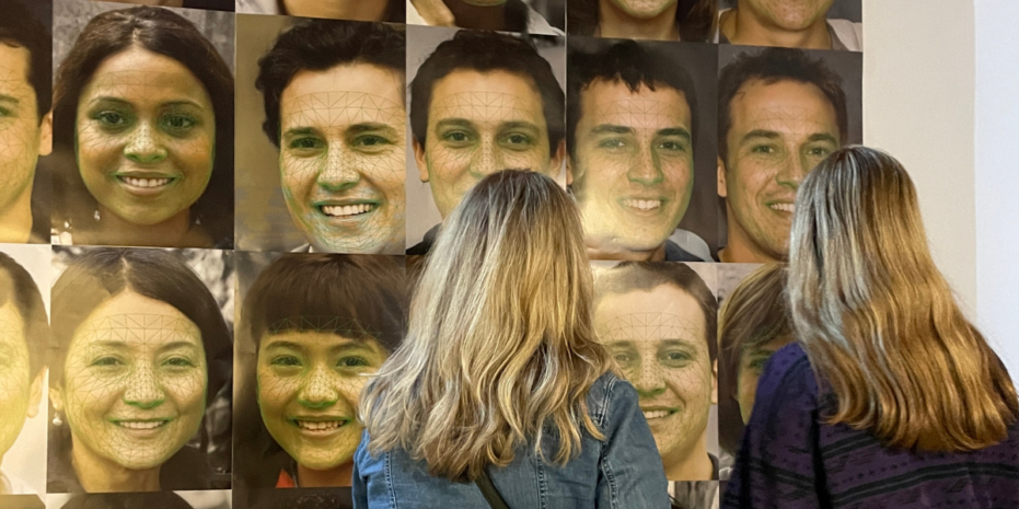 two women standing in front of a large wall display featuring multiple portrait photographs. Each portrait depicts a person's face with green geometric facial recognition mapping overlaid on the image, indicating the use of technology, likely AI, to analyze or track facial features. The women are observing the images closely, with their backs to the camera, engaging with the exhibit. The display seems to focus on AI or facial recognition technology, raising questions about identity, privacy, or the impact of AI on personal data. The setting appears to be a museum or gallery.