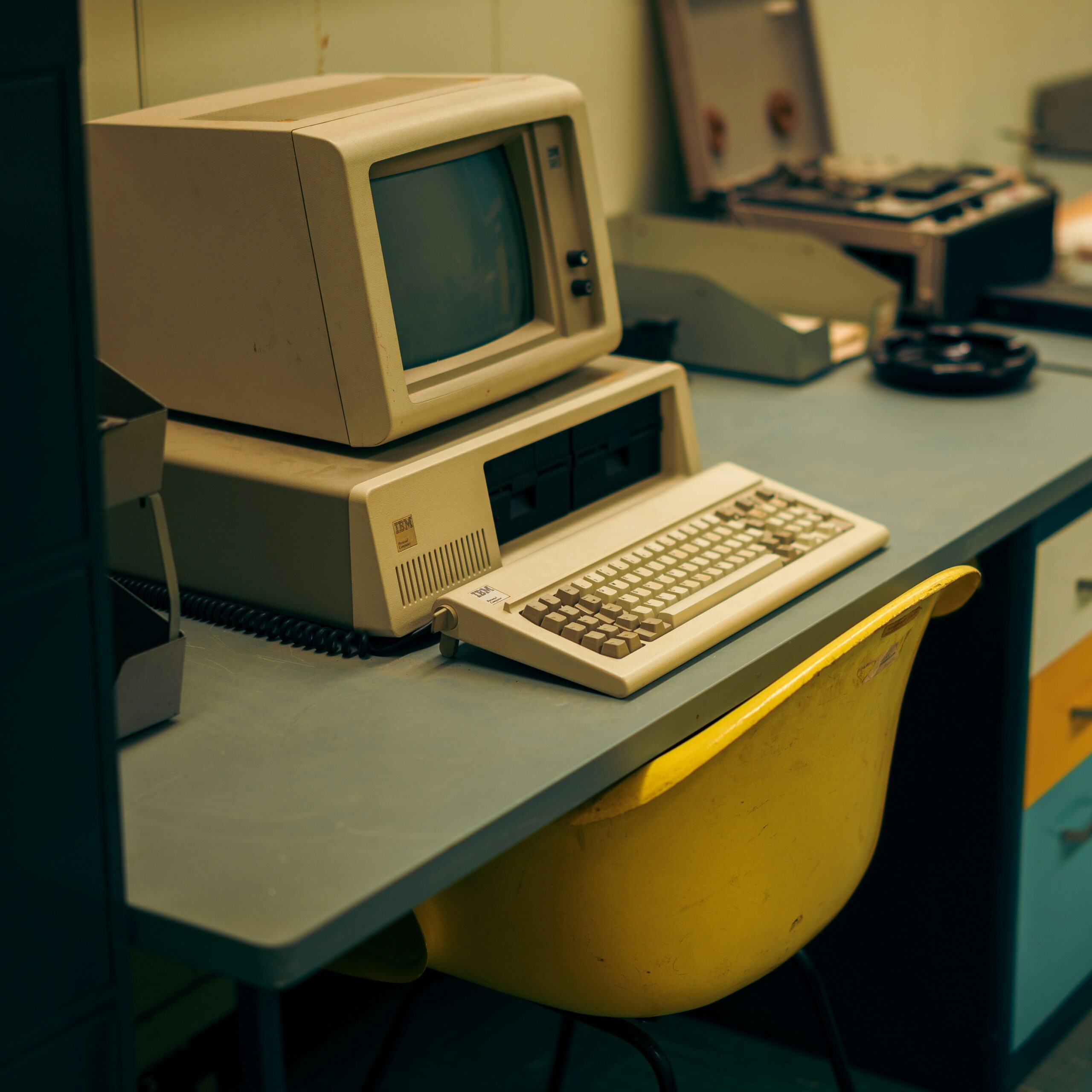 a vintage computer with a beige CRT monitor, floppy disk drives, and a classic keyboard sits on a muted blue desk with a coiled phone cable visible next to the keyboard. In front of the desk is a worn, yellow plastic chair. The background includes other retro office equipment, giving the space an old-fashioned, analog feel. The overall color scheme is subdued, evoking a nostalgic atmosphere of early computing days.