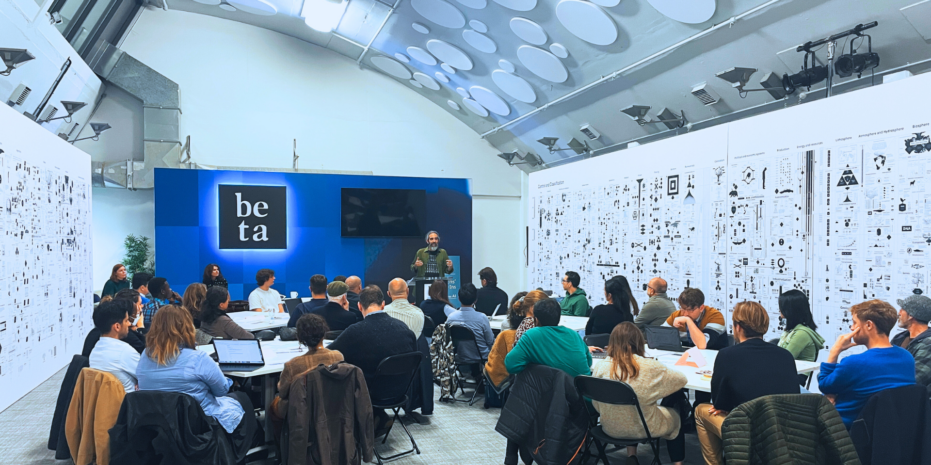 discussion event in progress at the Beta Festival. The room is filled with attendees seated at round tables, actively engaging with the speaker standing at the front. Large white panels line the walls, filled with intricate, black-and-white diagrams, charts, and illustrations, giving the setting a modern, data-focused feel. The focal point at the front of the room is a blue-lit wall with the Beta logo in white. The atmosphere is collaborative and intellectual, as participants listen attentively or discuss with one another.