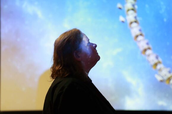 A person silhouetted in profile against a vivid background of a blue sky with clouds and a curving, futuristic structure, evoking a contemplative or futuristic atmosphere.