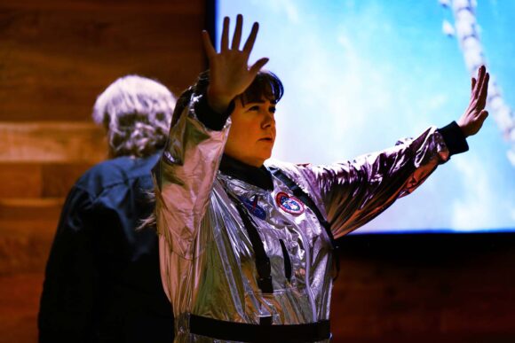 This image captures a moment from a performance. The person in a silver astronaut-style suit has their arms raised. The vibrant backdrop and dramatic lighting add to the dynamic and immersive atmosphere of the event.