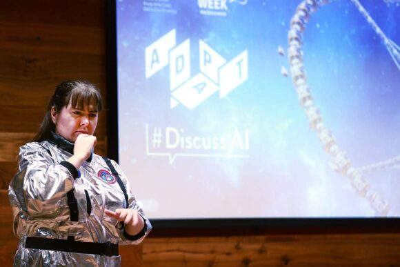 This image showcases a theatre in a silver astronaut-style suit performing through Irish Sign Language (ISL) in front of a backdrop displaying the logos of ADAPT and #DiscussAI. The event is part of a Science Week performance engaging with artificial intelligence and innovative discussions.