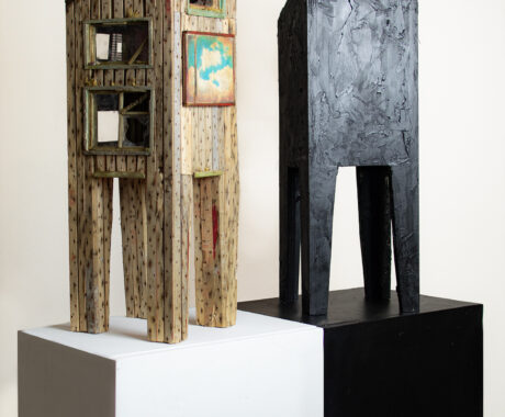 Two wooden sculptures on pedestals. The sculpture on the left is made of light-colored, weathered wood with visible nail holes, window-like openings, and a small painting of clouds framed on the front. The sculpture has a rustic, worn appearance. The sculpture on the right is taller and painted in solid black, with a textured surface that resembles darkened, charred wood. Both sculptures are rectangular with elongated legs, giving them a tower-like structure. The left sculpture stands on a white pedestal, and the right sculpture is on a black pedestal.