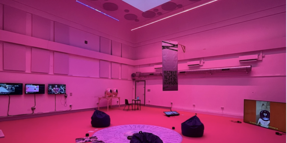 A modern, immersive installation space bathed in vibrant pink lighting. The room features a clean, minimalist design with bean bags arranged around a circular graphic on the floor, which appears to include text or diagrams. Several screens are mounted on the walls, displaying various visuals. A small table with equipment is positioned in the back, along with what looks like an artistic installation or sculpture. A vertical banner hangs from the ceiling, featuring a QR code. The atmosphere feels futuristic and engaging, inviting visitors to explore the technology and ideas presented in the space.