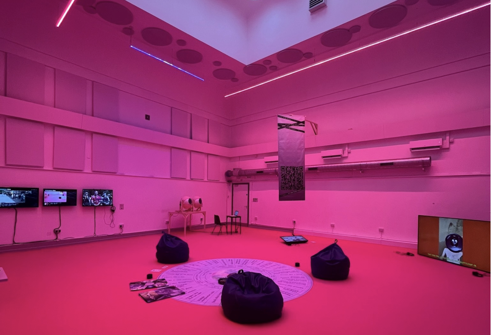 A modern, immersive installation space bathed in vibrant pink lighting. The room features a clean, minimalist design with bean bags arranged around a circular graphic on the floor, which appears to include text or diagrams. Several screens are mounted on the walls, displaying various visuals. A small table with equipment is positioned in the back, along with what looks like an artistic installation or sculpture. A vertical banner hangs from the ceiling, featuring a QR code. The atmosphere feels futuristic and engaging, inviting visitors to explore the technology and ideas presented in the space.