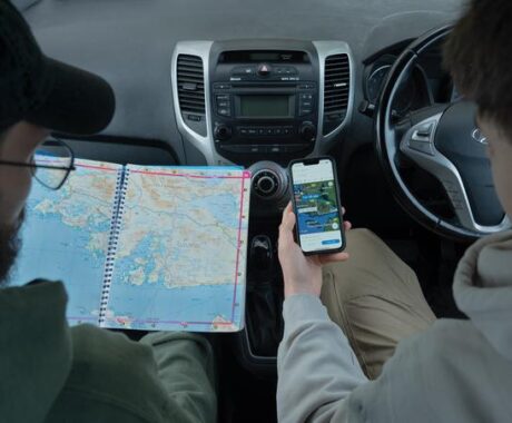 Two men are seated in the front seats of a car and the image is from the perspective as looking over the shoulders of the two individuals. The driver, on the right, holds a smartphone showing Google Maps with a route from Galway to Clifden, displaying the travel time and directions. The passenger, on the left, wears glasses and a dark green hoodie and holds a large, spiral-bound road atlas open to a page showing the west coast of Ireland. The car’s dashboard and controls are visible between them.