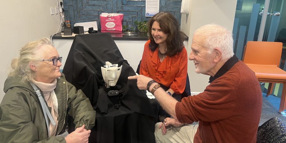 Three people—two women and a man—are seated around a robotic face covered in black fabric. The man is pointing at the robot while the others look on with interest. The setting appears to be an indoor public space with a modern interior design, including a blue-tiled wall, a countertop with a pink bag, and orange chairs in the background.